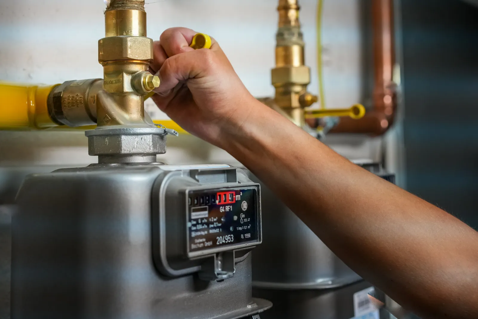 A hand turning the yellow valve above a gas meter, with the meter dial counting cubic metres of gas used